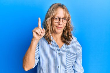 Orta yaşlı sarışın kadın, günlük kıyafetler ve gözlük takıyor. Bir numaralı parmağını gösteriyor ve işaret ediyor. Kendine güvenen ve mutlu bir şekilde gülümsüyor.. 