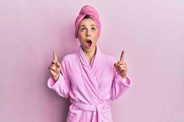 Duş başlığı ve bornoz takan genç sarışın kadın şaşırmış ve şaşırmış bir şekilde yukarı bakıyor ve parmaklarıyla işaret ediyor ve kollarını kaldırıyor.. 