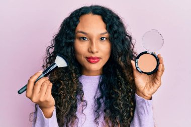 Makyaj fırçası tutan ve yüzünde ciddi bir ifadeyle rahatlamış genç Latin bir kadın. Basit ve doğal bir şekilde kameraya bakıyorsun.. 