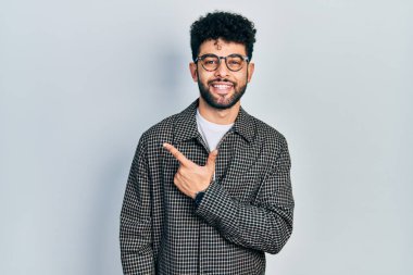 Sakallı, neşeli gözlükler takan genç bir Arap adam. Yüzünde mutlu ve doğal bir ifadeyle elleri ve parmaklarıyla yüzünü gösteren bir gülümseme. 
