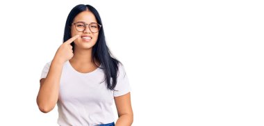 Genç, güzel Asyalı bir kız, günlük kıyafetler ve gözlük takıyor, el parmağı ve burunla işaret ediyor, neşeyle gülümsüyor. güzellik konsepti 