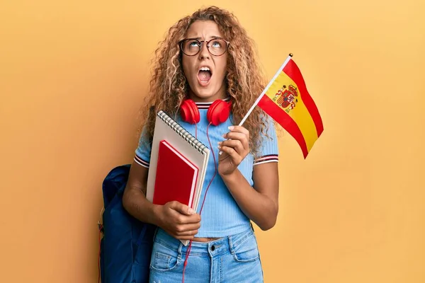 Beautiful caucasian teenager girl exchange student holding spanish flag angry and mad screaming frustrated and furious, shouting with anger looking up.