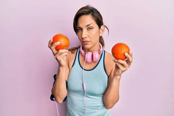 年轻的拉丁女人穿着运动服 用耳机 拿着橙子 疑神疑鬼 神经紧张 因为问题皱着眉头 消极的人 — 图库照片