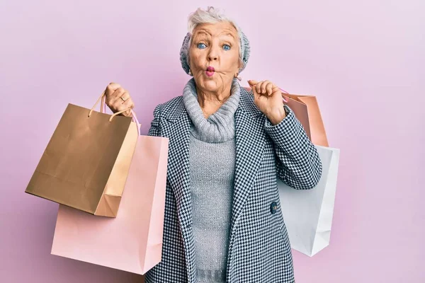 Senior grey-haired woman holding shopping bags making fish face with mouth and squinting eyes, crazy and comical.