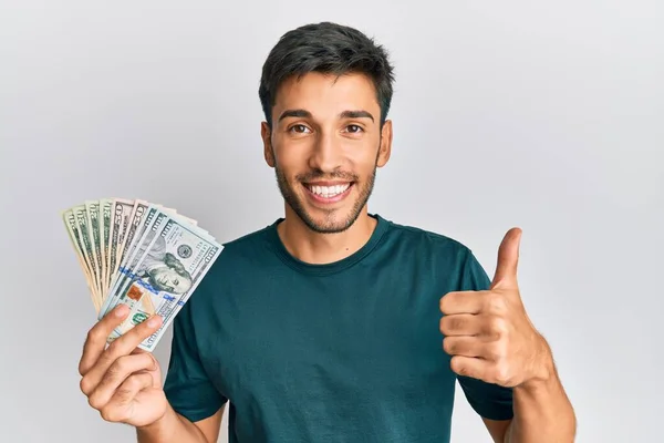 Joven Hombre Guapo Sosteniendo Dólares Sonriendo Feliz Positivo Pulgar Hacia —  Fotos de Stock