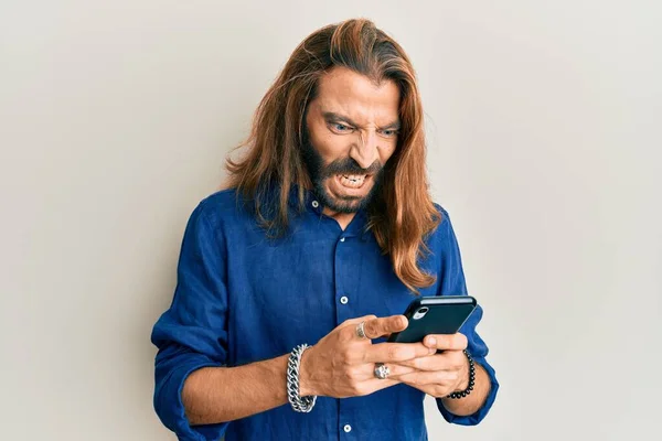 Attractive man with long hair and beard using smartphone angry and mad screaming frustrated and furious, shouting with anger. rage and aggressive concept.