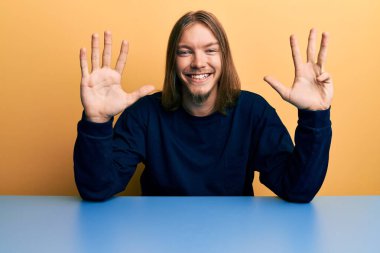 Uzun saçlı, yakışıklı beyaz bir adam masanın üzerine oturmuş dokuz numarayı gösterirken kendinden emin ve mutlu bir şekilde gülümsüyor.. 