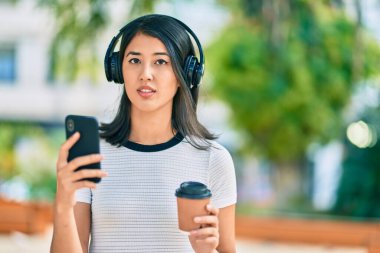 Genç İspanyol kadın kahve içiyor ve şehirde akıllı telefon ve kulaklık kullanıyor..