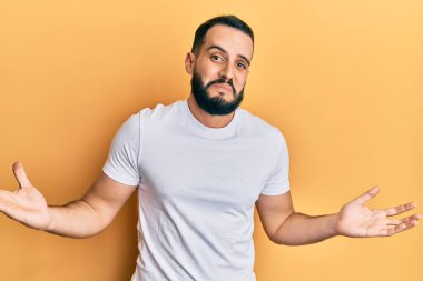 Sakallı, sıradan beyaz tişörtlü, kafası karışık, kolları ve elleri havada genç bir adam. şüphe kavramı. 