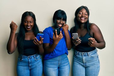 Akıllı telefon kullanan üç Afro-Amerikalı arkadaş çok mutlu ve heyecanlı bir şekilde kollarını kaldırıp, gülümseyerek ve başarı çığlıkları atarak zafer jesti yapıyorlar. kutlama konsepti. 