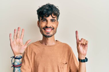 Latin asıllı genç adam gündelik giysiler giyip altı numaralı parmaklarıyla işaret ederken kendinden emin ve mutlu bir şekilde gülümsüyor.. 