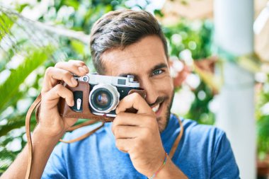 Genç yakışıklı fotoğrafçı parkta geçmişe dönük kamera kullanarak mutlu bir şekilde gülümsüyor.