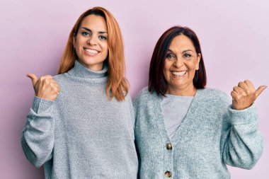 Latin anne ve kız, gündelik giysiler giyip elleri ve başparmakları havada, kendinden emin bir şekilde arkasını gösteriyorlar. 