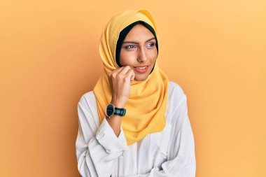 Genç esmer Arap kadın geleneksel İslami başörtüsü takıyor ve elleri çenesinde soru sormayı, düşünceli ifadeyi düşünüyor. Gülümseyen ve düşünceli bir yüz. şüphe kavramı. 