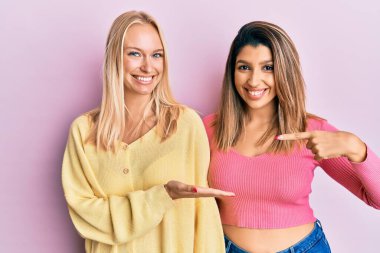 Pembe arka planda yan yana duran iki arkadaş kameraya hayret ediyor ve gülümsüyor.. 