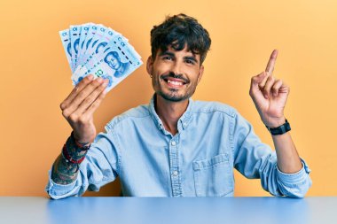 Genç İspanyol adam Yuan Çin banknotlarını tutuyor masaya oturmuş gülümsüyor mutlu bir şekilde el ve parmak kenarı gösteriyor. 