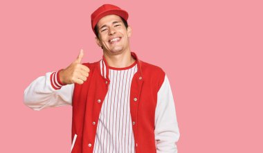 Beyzbol üniforması giyen yakışıklı beyaz adam mutlu bir el hareketi yapıyor. Kameraya bakıp başarıyı gösteren ifade. 