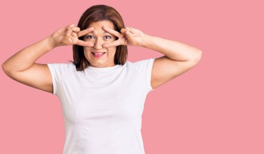 Orta yaşlı beyaz tişört giyen Latin bir kadın barış sembolü yapıyor. Yüzünde parmakları var. Gülümsüyor, zaferini gösteriyor. 