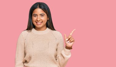 Kışlık yün kazak giyen, yüzünde kocaman bir gülümseme olan genç Latin kız, kameraya el ile işaret ediyor.. 