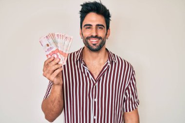 Sakallı yakışıklı, Kolombiya pezosu tutan yakışıklı. Olumlu ve mutlu görünüyor. Kendine güvenen bir gülümsemeyle dişlerini gösteriyor. 