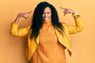 Orta yaşlı Afro-Amerikalı kadın yün kazak ve deri ceket giyiyor. İki eliyle başını gösteren bir gülümseme. Harika bir fikir ya da düşünce. 