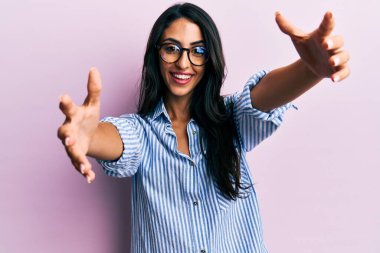 Güzel İspanyol kadın, günlük kıyafetler ve gözlük takmış kameraya bakıyor sarılmak için kollarını açmış gülümsüyor. Mutluluğu kucaklayan neşeli bir ifade.. 