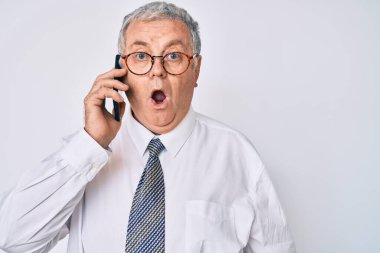 Yaşlı, gri saçlı, iş kıyafetleri giyen, akıllı telefonda konuşan, ağzı açık, korkmuş ve şaşırmış bir adam. 