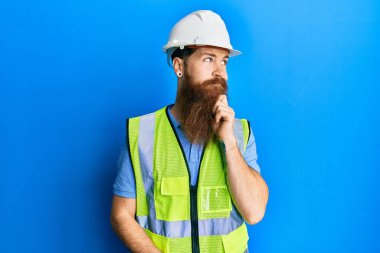 Uzun sakallı, güvenlik kaskı takan, eli çenesinde yansıtıcı ceketli, soru soran, düşünceli bir adam. Düşünceli bir yüzle gülümsüyor. şüphe kavramı. 