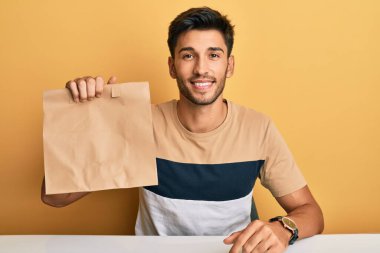 Young handsome man holding take away paper bag looking positive and happy standing and smiling with a confident smile showing teeth 