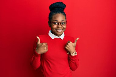 Genç Afro-Amerikalı kadın günlük kıyafetler ve gözlük takıyor. Başarı işareti el hareketleriyle olumlu jest yapıyor. Gülümsüyor ve mutlu. Neşeli ifade ve kazanan jest. 