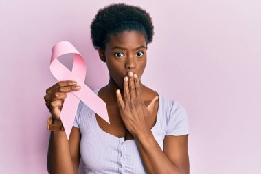 Genç Afro-Amerikan kızı elinde pembe kanser kurdelesi tutarak ağzını eliyle kapatmış, şok olmuş ve hata yapmaktan korkan. şaşırmış ifade 