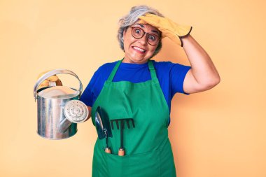 Bahçıvan önlüğü ve eldiven giyen yaşlı, beyaz saçlı bir kadın. Sulama kabı taşıyor. Elleri başının üstünde, şaşırmış ve kızgın bir yüz. 