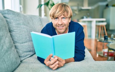 Orta yaşlı, yakışıklı, beyaz bir adam evde kitap okuyor.