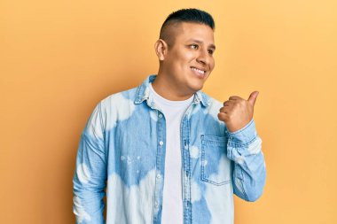 Gündelik giysiler giymiş genç Latin bir adam başparmağını yana kaldırmış, ağzı açık gülümsüyor. 