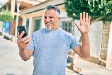 Orta yaşlı, ispanyol, gri saçlı, gülümseyen, şehirde akıllı telefon kullanarak video araması yapan bir adam..