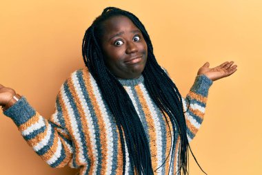 Saçları örgülü genç siyah bir kadın. Sıradan bir kazak giyiyor. Kolları ve elleri havada, kafası karışık bir ifade takınıyor. şüphe kavramı. 