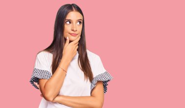 Elleri çenesinde gündelik kıyafetler giyen genç İspanyol kadın soru sormayı düşünüyor, düşünceli bir ifade. Düşünceli bir yüzle gülümsüyor. şüphe kavramı. 