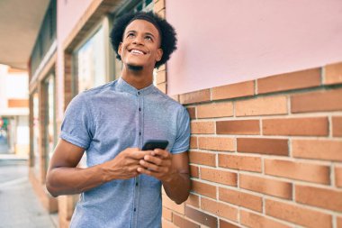 Genç Afrikalı Amerikalı adam şehirde akıllı telefon kullanarak mutlu bir şekilde gülümsüyor..