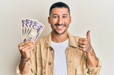 Japon yen banknotları tutan yakışıklı adam mutlu ve olumlu gülümsüyor, başparmağıyla mükemmel ve onay işareti yapıyor. 