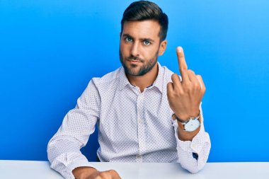 Handsome hispanic man wearing business clothes sitting on the table showing middle finger, impolite and rude fuck off expression 