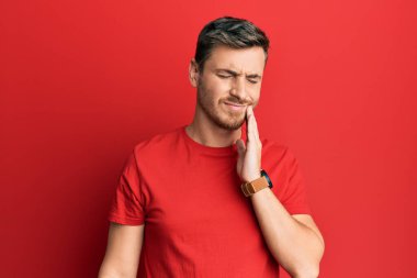 Yakışıklı beyaz bir adam dişleri ağrıdığı ya da dişlerinde hastalık olduğu için elleri ve elleri birbirine değen gündelik kırmızı tişört giyiyor. dişçi 