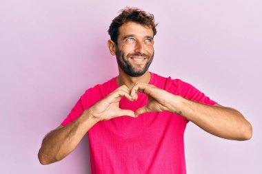 Sakallı yakışıklı adam, elleri şekilli kalp sembolü yapıyor. Gülümsüyor ve başka tarafa bakıyor.. 