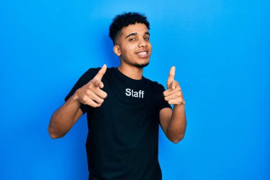 Genç, Afro-Amerikalı, personel tişörtü giymiş, mutlu ve komik yüzüyle kamerayı işaret eden bir adam. iyi enerji ve titreşimler. 