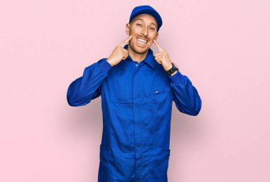 Sakallı, inşaatçı tulumu giyen, ağzı açık gülümseyen, parmakları işaret eden ve neşeli gülümsemeye zorlayan kel bir adam. 