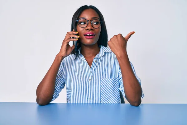 Joven Mujer Afroamericana Hablando Teléfono Inteligente Sentado Mesa Apuntando Pulgar —  Fotos de Stock