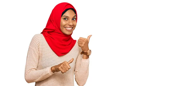 手と親指を上げて後ろを指差す伝統的なイスラムのヒジャーブスカーフを身に着けている若いアフリカ系アメリカ人女性は 自信を持って笑って — ストック写真