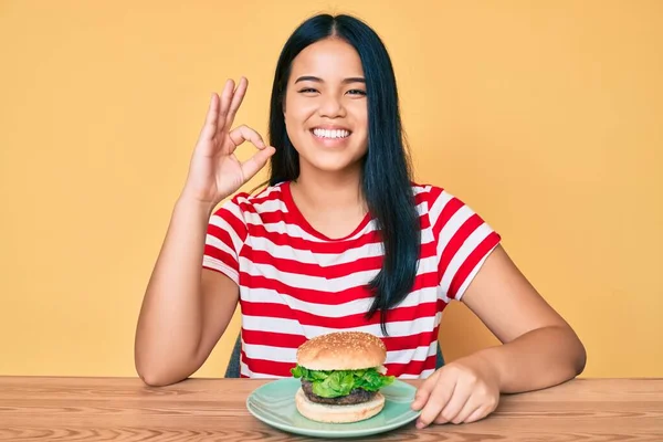 年轻的亚洲女孩吃着美味的经典汉堡包 手拿着手签 微笑着表示友善的手势 是极好的象征 — 图库照片