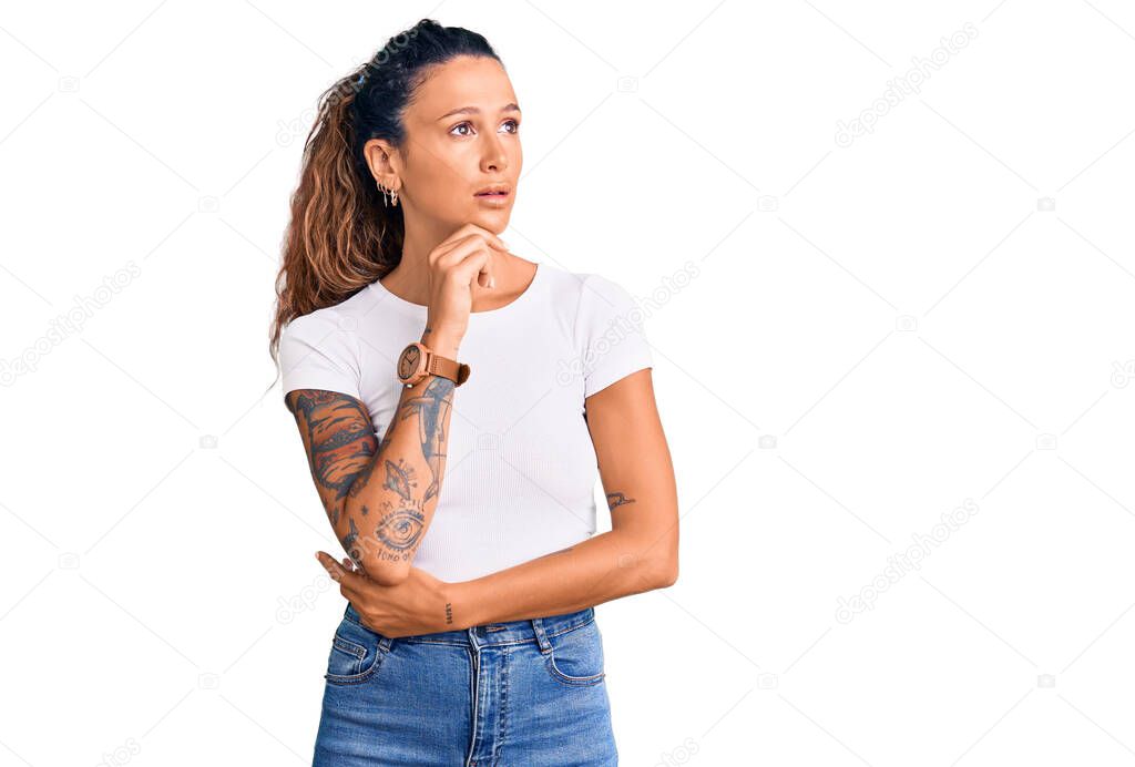 Young hispanic woman with tattoo wearing casual white tshirt with hand on chin thinking about question, pensive expression. smiling with thoughtful face. doubt concept. 
