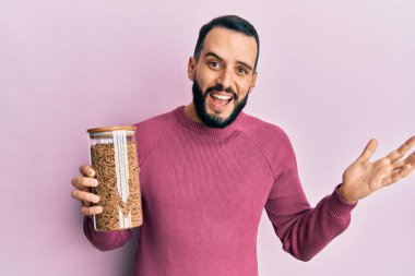 Sakallı genç bir adam sağlıklı tahıl gevreği kavanozunu tutarak başarıyı mutlu bir gülümsemeyle ve kazanan yüz ifadesiyle kutluyor. 