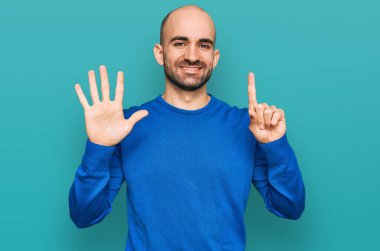 Latin asıllı genç adam gündelik giysiler giyip altı numaralı parmaklarıyla işaret ederken kendinden emin ve mutlu bir şekilde gülümsüyor.. 